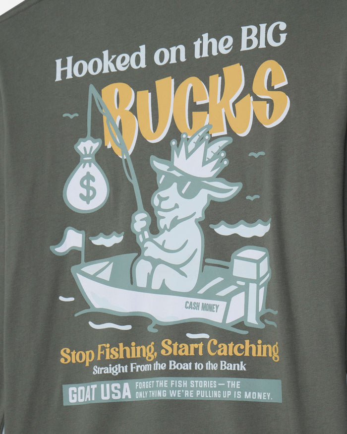 Close-up of a dark olive green t-shirt featuring a graphic print with a cartoon goat in a boat holding a fishing rod and money bag, with text reading 'Hooked on the BIG Bucks' and 'Stop Fishing, Start Catching'.