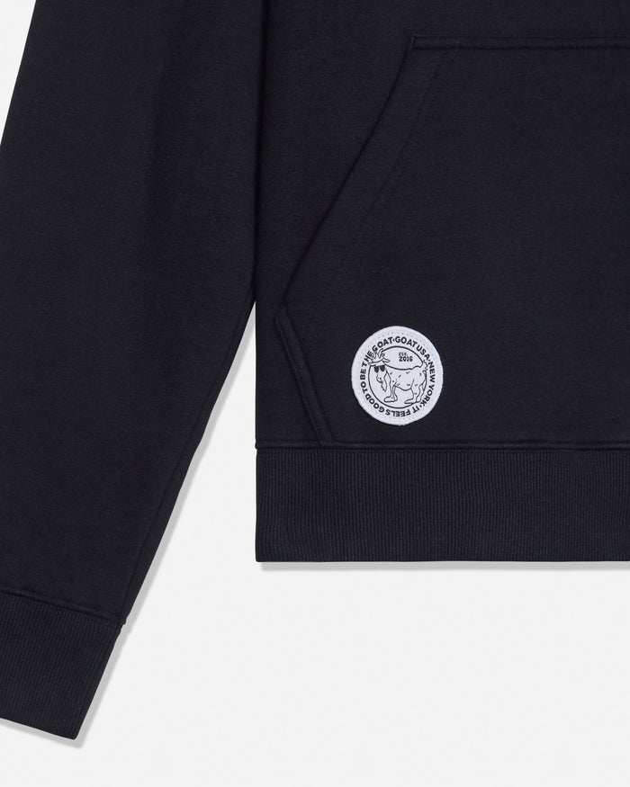 Close-up of a black sweatshirt sleeve with a circular white embroidered logo patch on the upper cuff, shown against a light gray background.