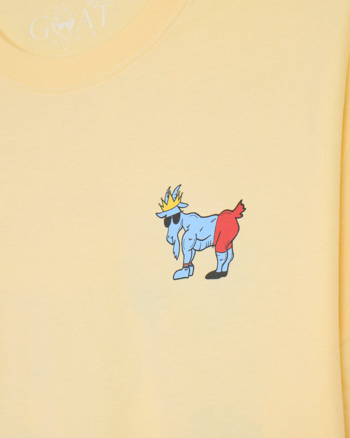 Close-up of a pale yellow T-shirt with a small cartoon goat graphic in the center wearing a gold crown, black sunglasses, and red shorts.