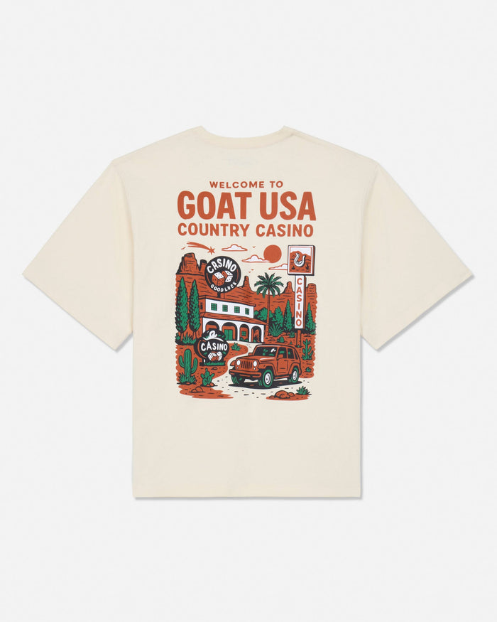 An off-white short-sleeve t-shirt with a large graphic print on the back featuring the text 'WELCOME TO GOAT USA COUNTRY CASINO' and an illustration of a red car, casino building, and desert scenery.