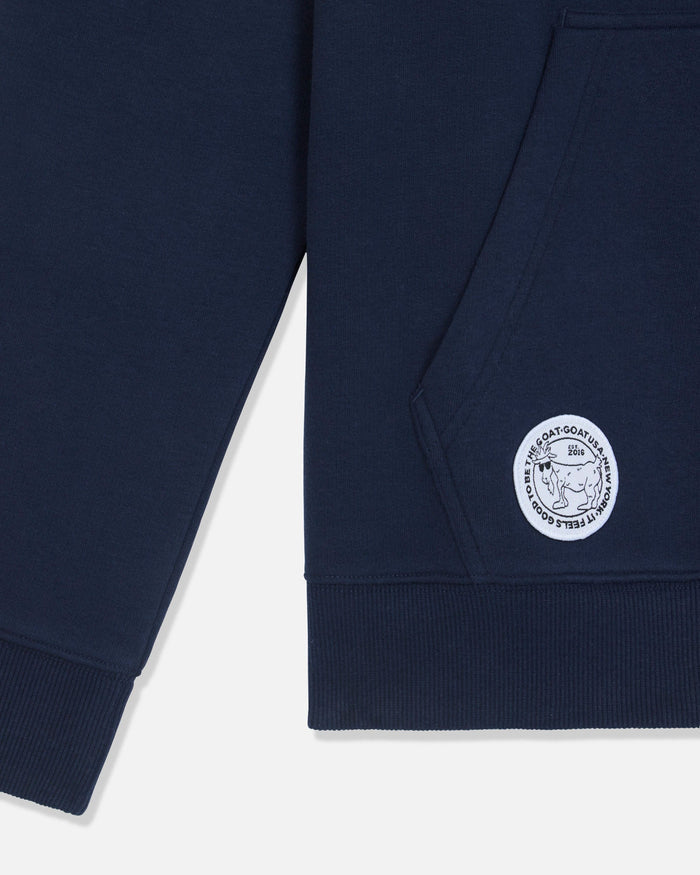 Close-up of a navy blue sweatshirt showing the sleeve, ribbed hem, and a circular white embroidered logo patch on the lower front.