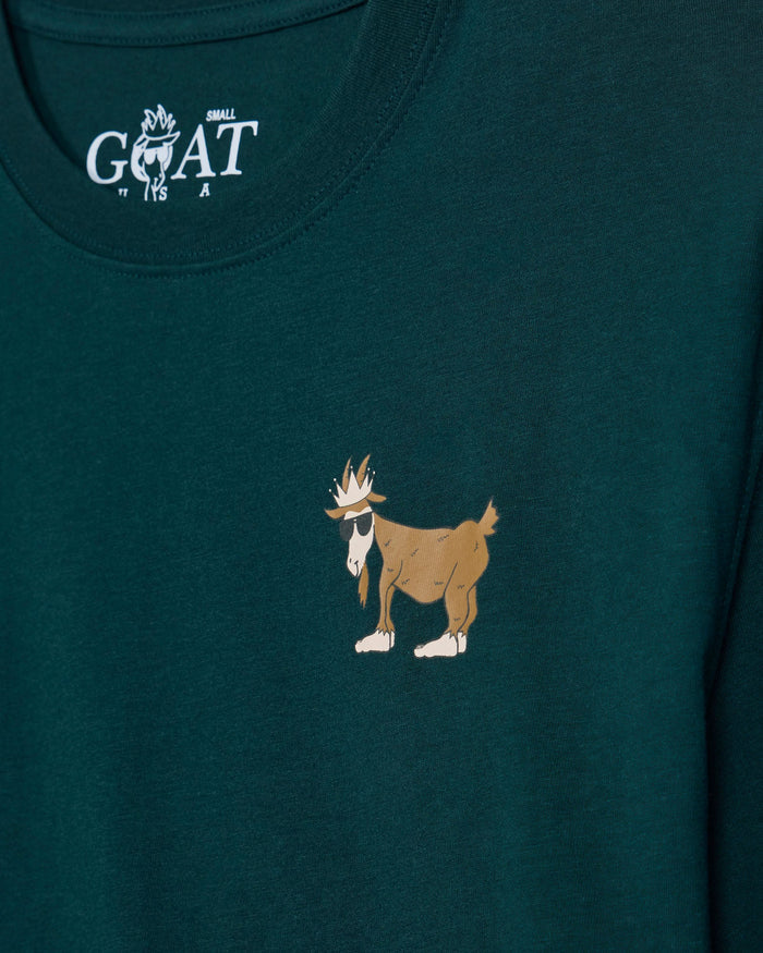 Close-up of a dark teal-green t-shirt featuring a small brown and white cartoon goat graphic on the center chest, with a white 'GOAT' brand logo printed inside the collar.
