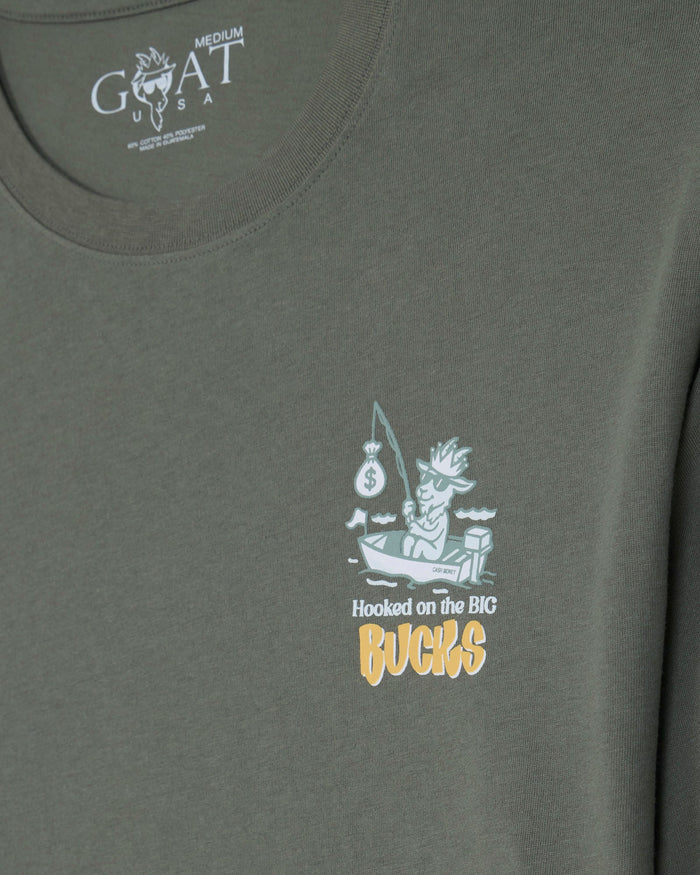 Close-up of a medium gray-green t-shirt featuring a small graphic on the left chest of a cartoon fisherman in a boat with a fishing rod and the text 'Hooked on the BIG Bucks' in white and yellow. The inside collar shows the Goat USA brand logo.