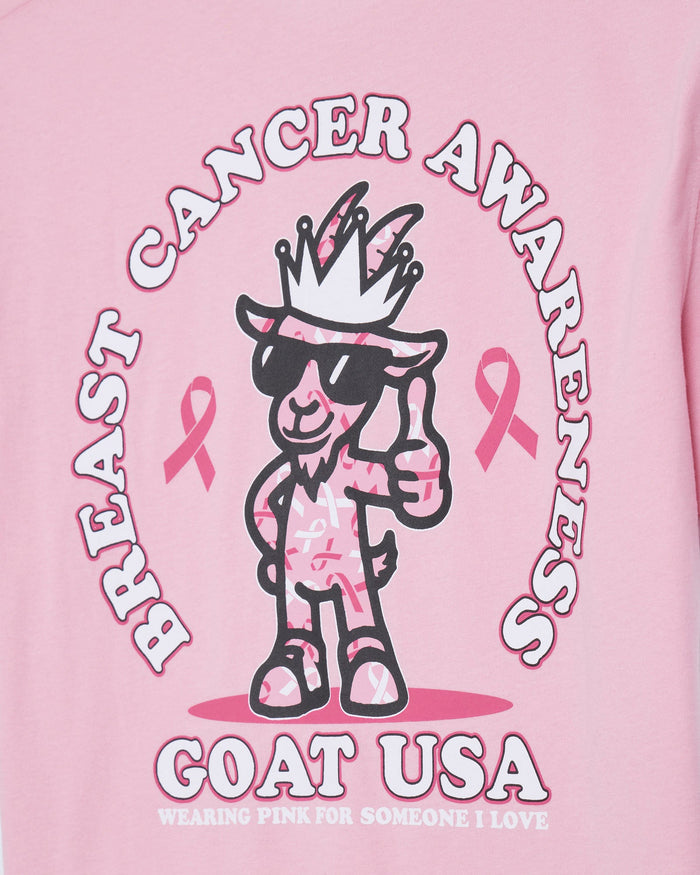 A close-up of a pink graphic t-shirt featuring a cartoon goat character wearing sunglasses and a crown, surrounded by the text 'BREAST CANCER AWARENESS' and 'GOAT USA'.