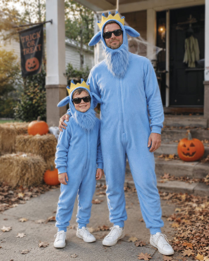 A man and a child wearing matching light blue goat onesies with yellow crown headpieces, standing on a front porch decorated for Halloween with pumpkins, hay bales, and spiderwebs.