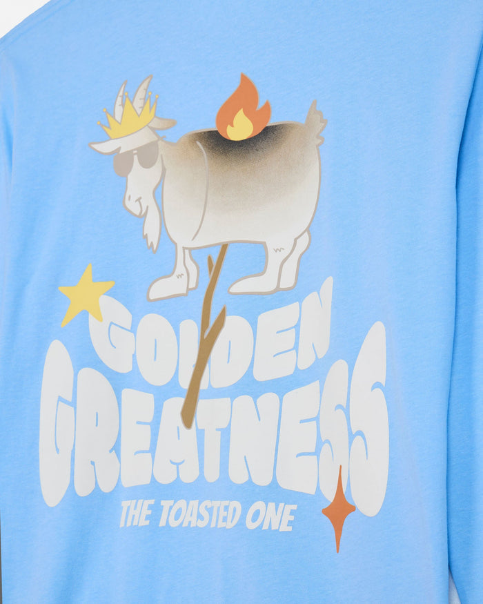 A close-up of a light blue long-sleeve shirt featuring a graphic print of a goat wearing a golden crown and sunglasses, with a flame on its back, and the text 'GOLDEN GREATNESS THE TOASTED ONE'.