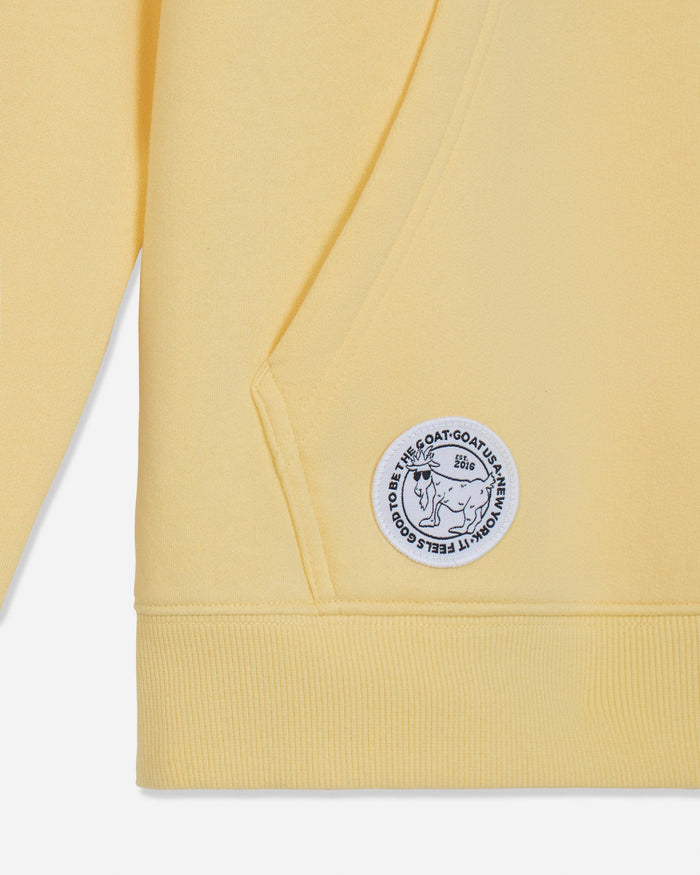Close-up of a pale yellow sweatshirt showing the ribbed cuff, shoulder seam, and a circular white patch with black text and a goat illustration on the upper sleeve.