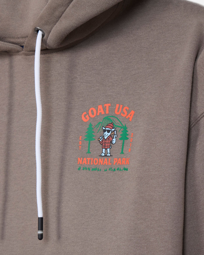 Close-up of a taupe-brown hooded sweatshirt featuring a white drawstring and a colorful chest graphic that reads 'GOAT USA NATIONAL PARK' with a cartoon goat in a plaid shirt and trees.