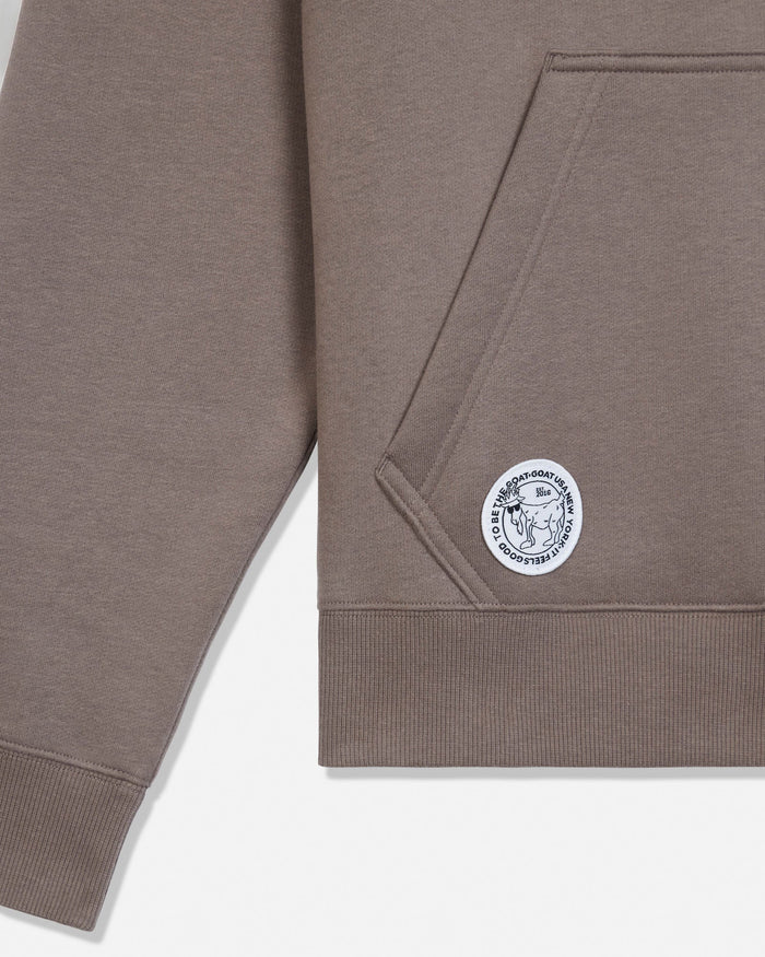 Close-up of a taupe-brown pullover hoodie showing the sleeve, pocket seam, ribbed hem, and a circular white embroidered logo patch on the lower front.