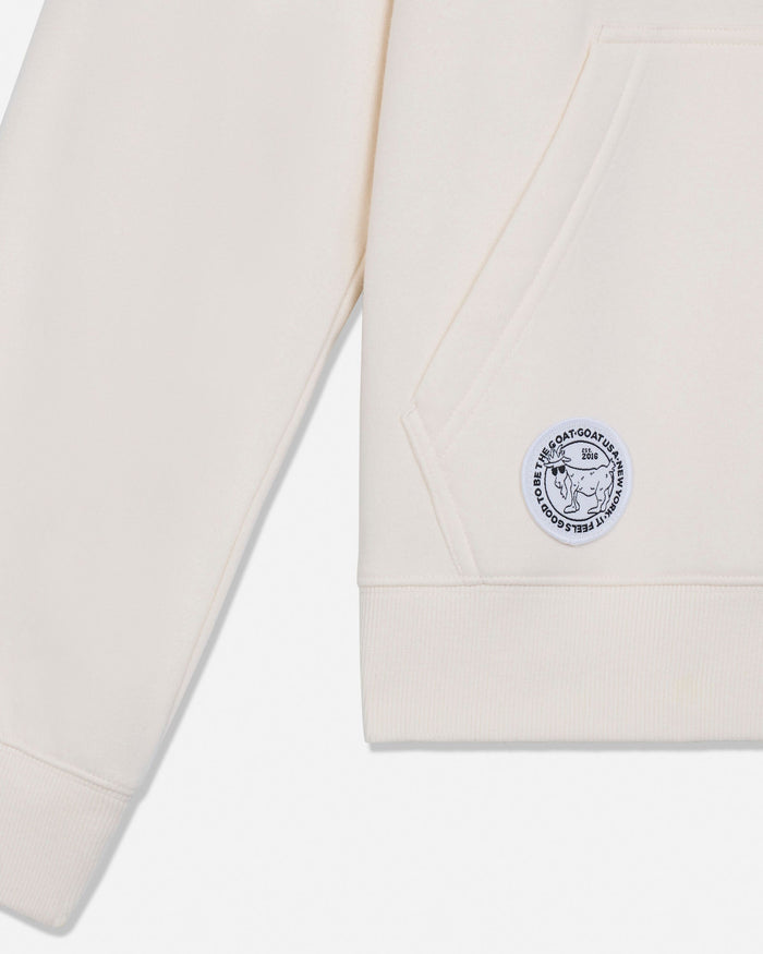 Close-up of a cream-colored sweatshirt showing the sleeve, pocket seam, ribbed hem, and a circular embroidered logo patch on the pocket.