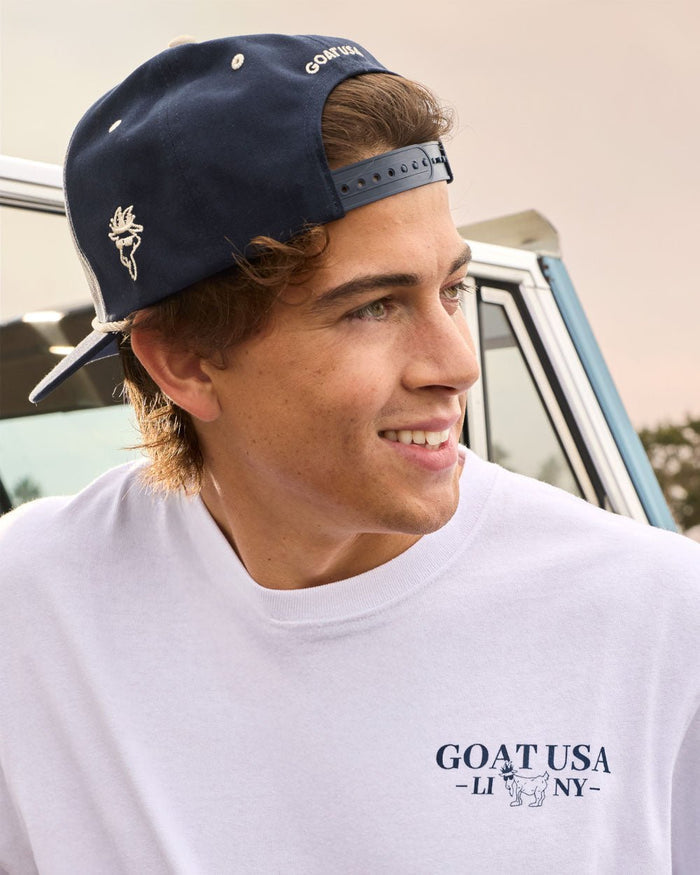 Person wearing a navy cap and white t-shirt with 'GOAT USA' logo, standing in front of a vehicle.