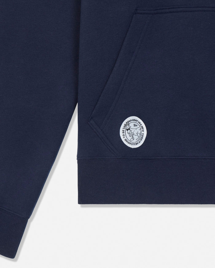 Close-up of a navy blue sweatshirt showing the sleeve cuff, ribbed hem, front pocket, and a circular embroidered logo patch on the chest.