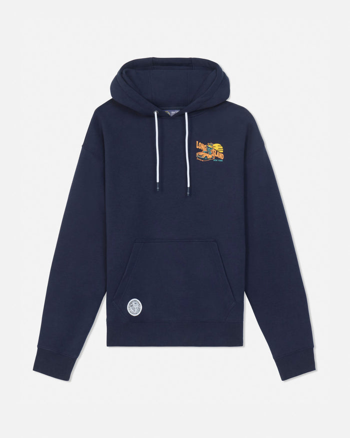 A navy blue pullover hoodie with white drawstrings, featuring a small yellow and orange graphic on the left chest and a circular white patch on the lower left front pocket, displayed against a light gray background.