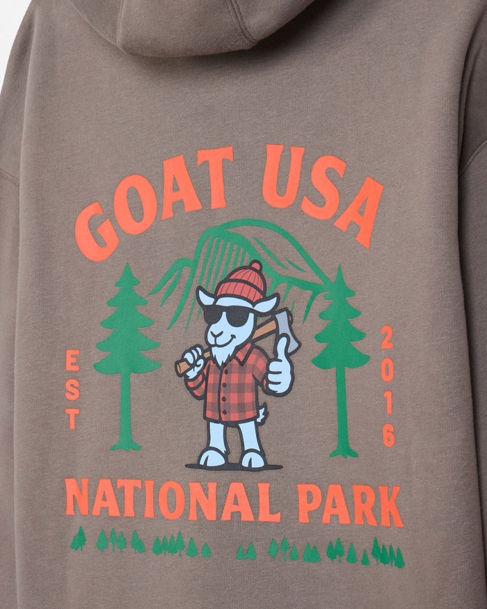 Close-up of the back of a taupe hoodie featuring a colorful graphic print with the text 'GOAT USA NATIONAL PARK', green pine trees, a cartoon goat in a red plaid shirt holding an axe, and 'EST 2016' printed vertically.