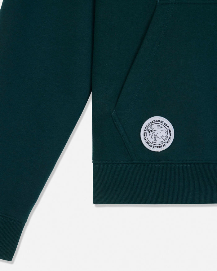 Close-up of a dark green sweatshirt sleeve and ribbed hem, featuring a circular white embroidered logo patch on the lower sleeve.
