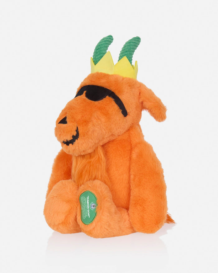 An orange plush goat toy with a yellow and green crown, black embroidered eyes and mouth, and a green circular tag on its front paw, photographed on a clean white background.