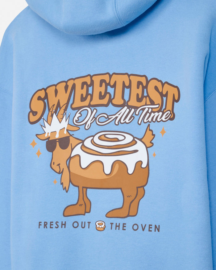 A close-up of a light blue hoodie showing a colorful graphic print on the front. The graphic features a cartoon moose wearing sunglasses and a crown, standing next to a cinnamon roll with white icing, with the text 'SWEETEST Of All Time' and 'FRESH OUT THE OVEN'.