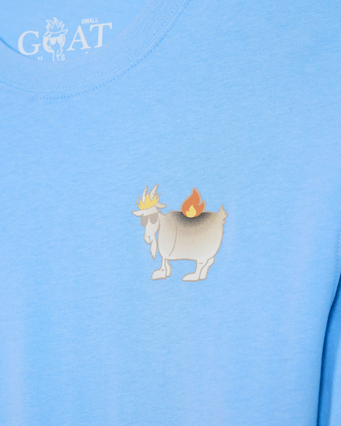 A close-up of a light blue t-shirt featuring a small cartoon graphic of a white goat with a yellow crown and a small orange fire on its back, centered on the chest. The brand name 'GOAT USA' is printed in white near the collar.