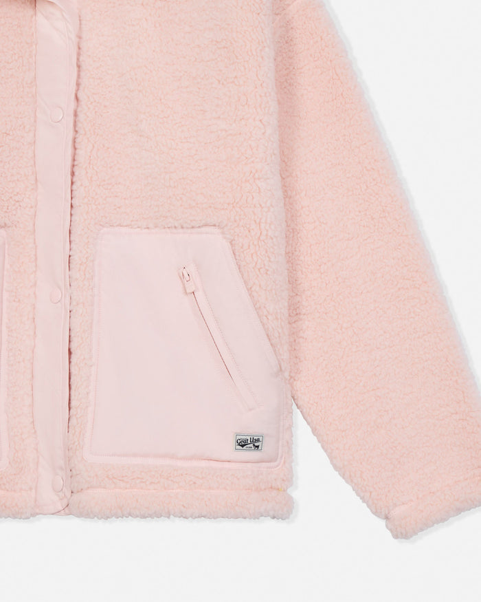 Close-up of a light pink sherpa fleece jacket with a zippered front pocket and a small metallic logo badge near the hem.