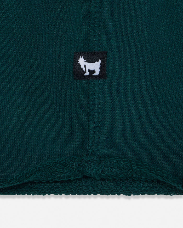 Close-up of a dark teal knit garment showing a small black square patch with a white goat logo, likely indicating the brand GOAT USA.